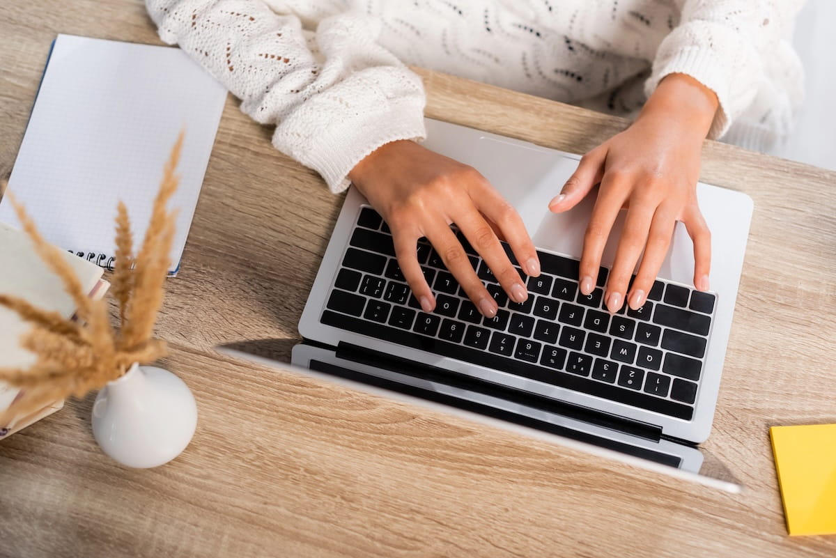 An overhead shot of a local newsletter creator's hands typing on a silver laptop at a wooden desk. To the left sits a blank white notepad and a small white vase containing dried pampas grass, while a single yellow sticky note is visible on the bottom right corner of the desk. The creator is wearing a white, textured knit sweater.