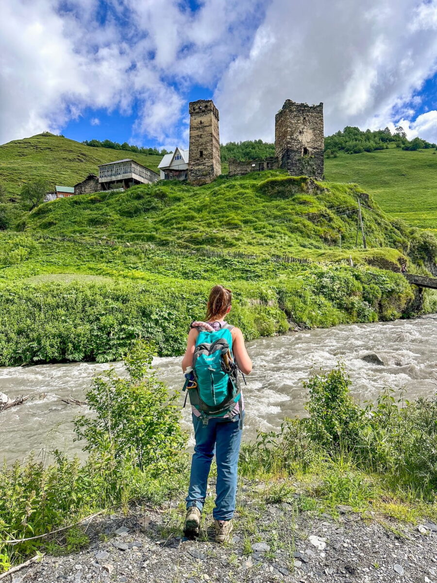 A female hiker from behind wearing a teal G4Free 35L Day Pack and blue hiking pants stands on the edge of a rushing mountain river, looking across at historic medieval stone defense towers sitting atop a lush green hill in Svaneti, Georgia.
