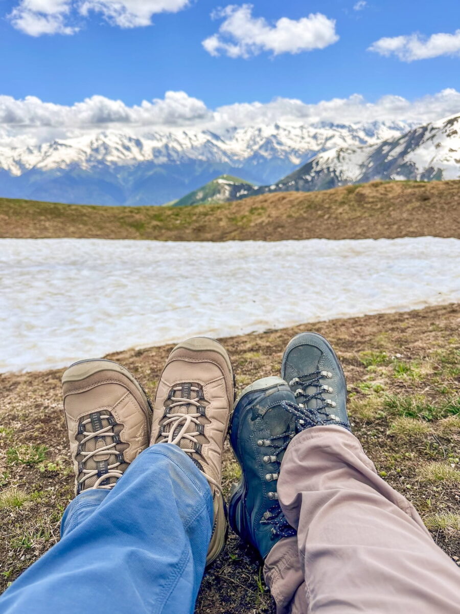 A first-person perspective looking down at two pairs of sturdy waterproof hiking boots, including beige KEEN waterproof boots, resting on a dirt bank overlooking a white alpine snowfield and the majestic, rugged snow-capped peaks of the Caucasus Mountains in Svaneti, Georgia.