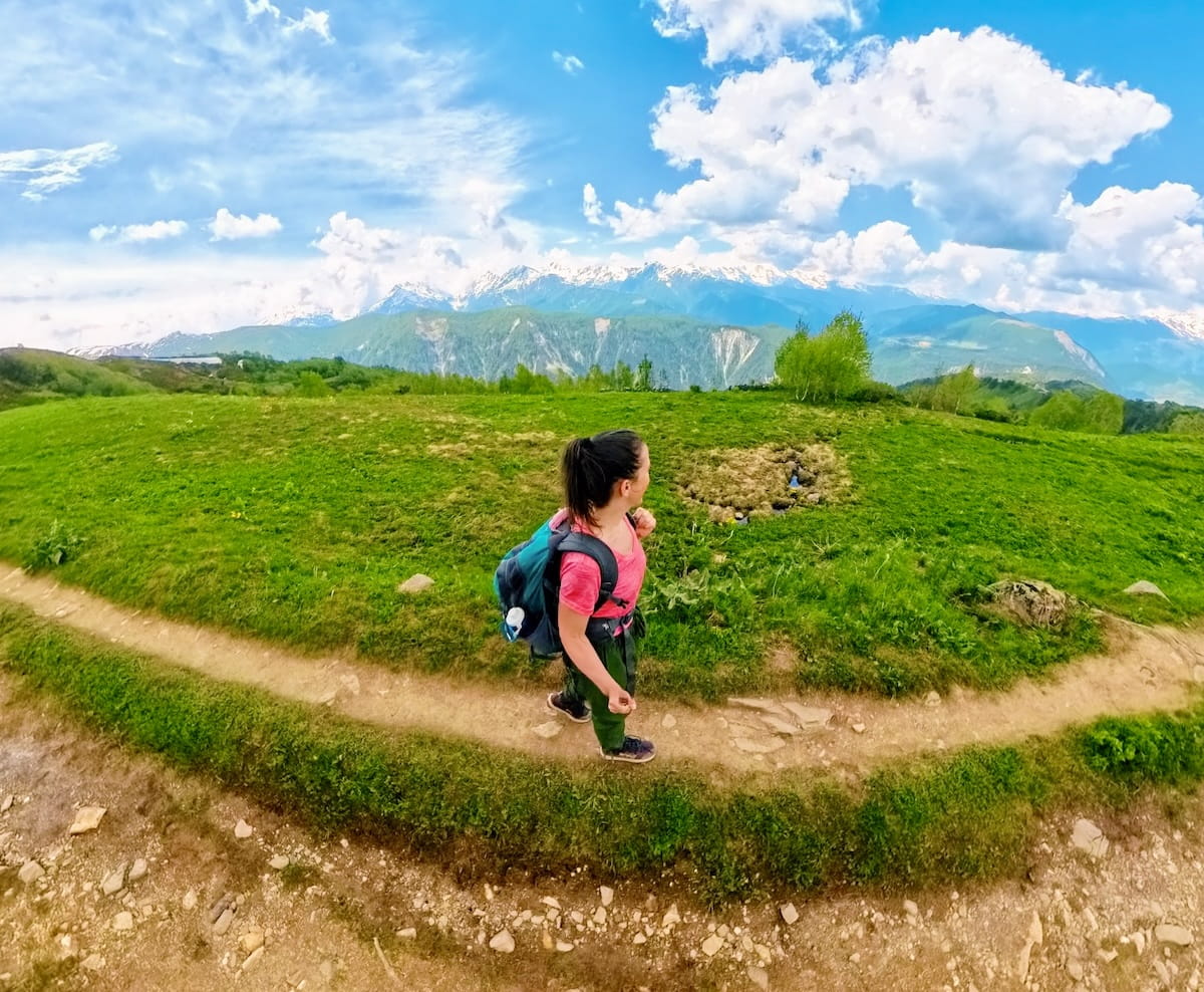 A high-angle 360 shot of a female hiker wearing a pink shirt, green pants, and a blue backpack as she walks along a winding dirt path cutting through a vibrant green alpine meadow, with the vast, snow-capped Caucasus Mountain range stretching across the horizon under a cloudy blue sky in Svaneti, Georgia.