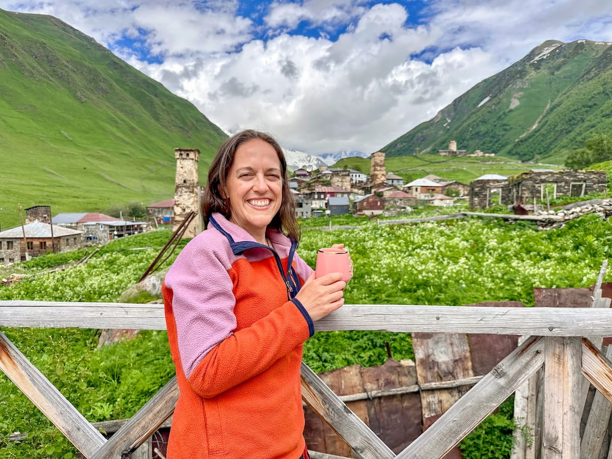 A smiling female hiker wearing a colorful orange and pink Cotopaxi fleece jacket stands on a wooden balcony holding a pink mug, overlooking a historic mountain village with stone defense towers nestled in a lush green valley beneath a cloudy sky in Svaneti, Georgia.