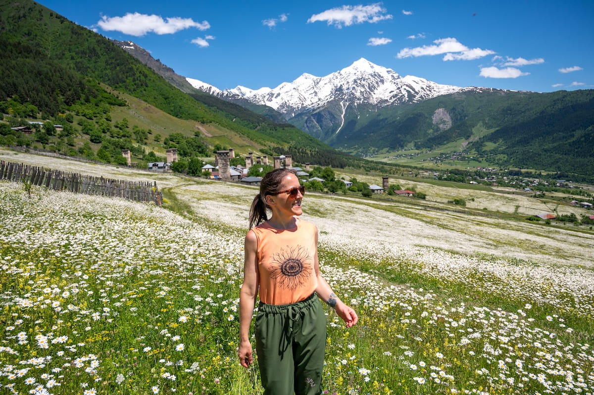 A smiling female hiker wearing brown Goodr polarized sunglasses, an orange tank top with a sunflower graphic, and green hiking pants stands in a massive field of white and yellow daisy wildflowers, with historic Svaneti stone defense towers and a towering, snow-capped Mount Tetnuldi rising under a blue sky in Georgia.