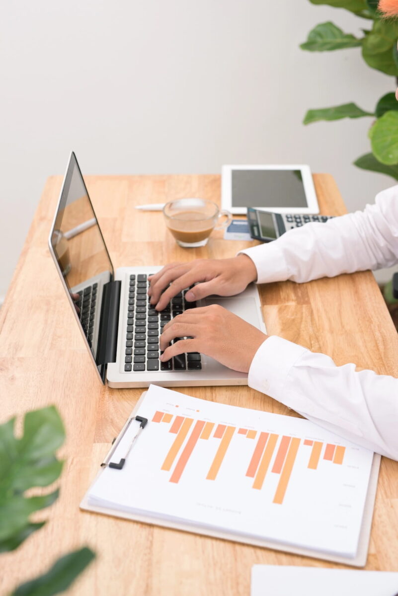 A top-angle shot of a blogger in a white button-down shirt typing on a laptop at a wooden desk. A clipboard with an orange bar chart, a cup of coffee, a calculator, and a tablet are also visible on the workspace.