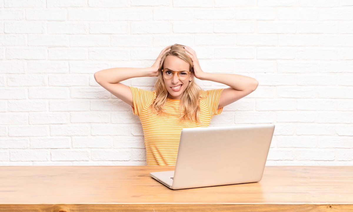 A blonde woman blogger wearing glasses and a yellow striped t-shirt sits at a wooden desk behind a laptop, holding her head in frustration against a white brick wall backdrop.