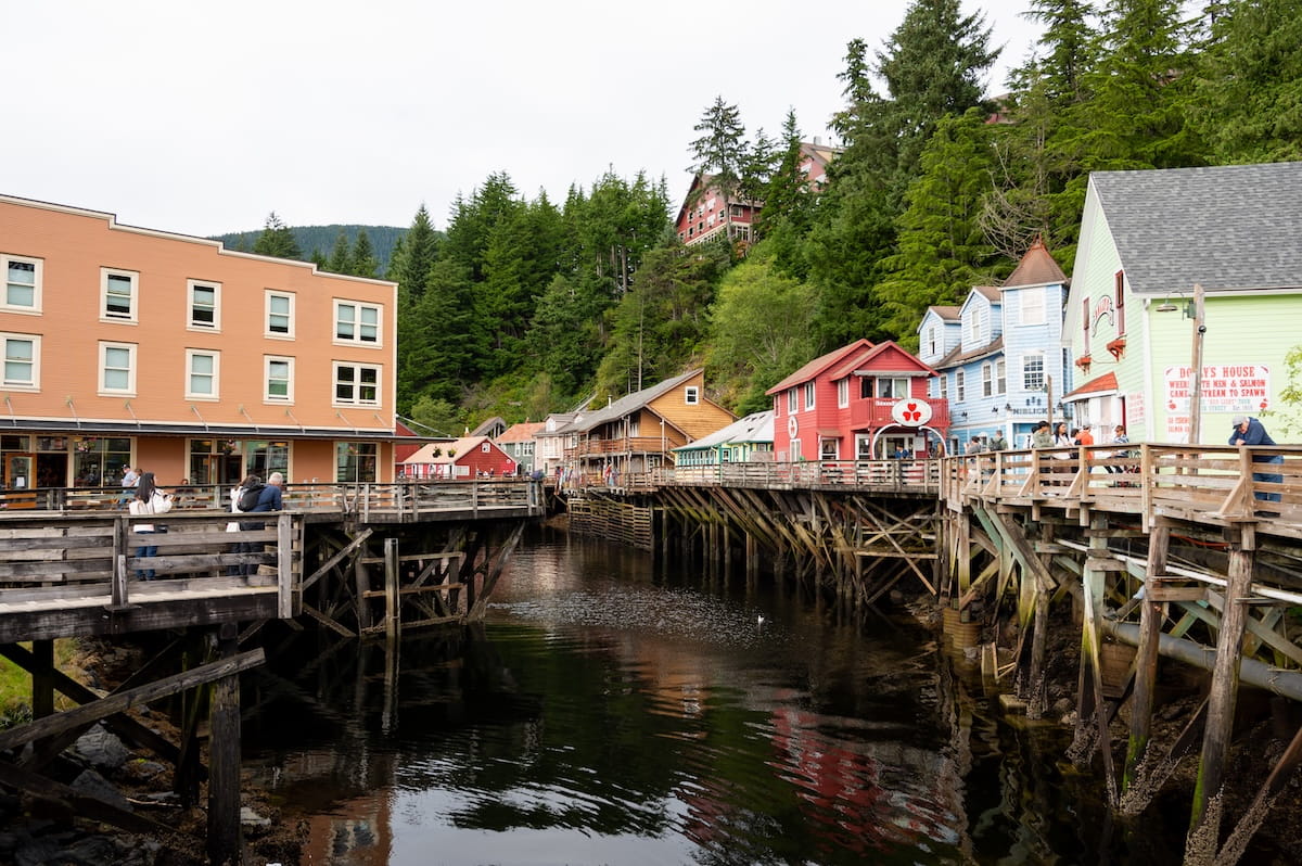 Colorful historic buildings on wooden stilts line the boardwalk of Creek Street in Ketchikan, Alaska, with their reflections cast on the calm water of the creek below and a dense green forest rising on the hillside in the background.