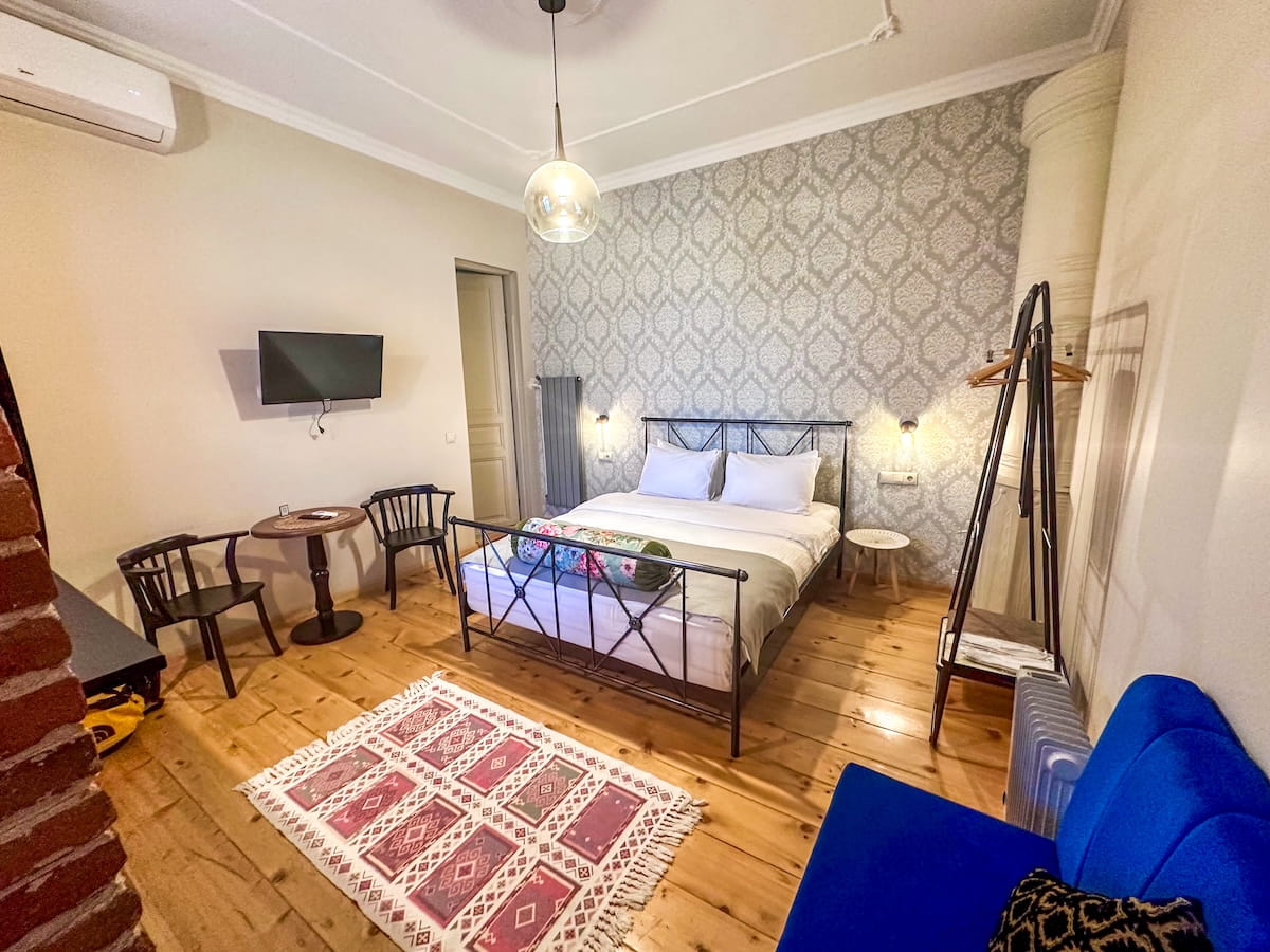 A boutique hotel room at Arts Hotel Tbilisi, featuring a wrought iron double bed against a grey damask patterned accent wall, light hardwood floors, a traditional patterned area rug, a small seating area with black wooden chairs, and a bright blue sofa chair in the foreground.