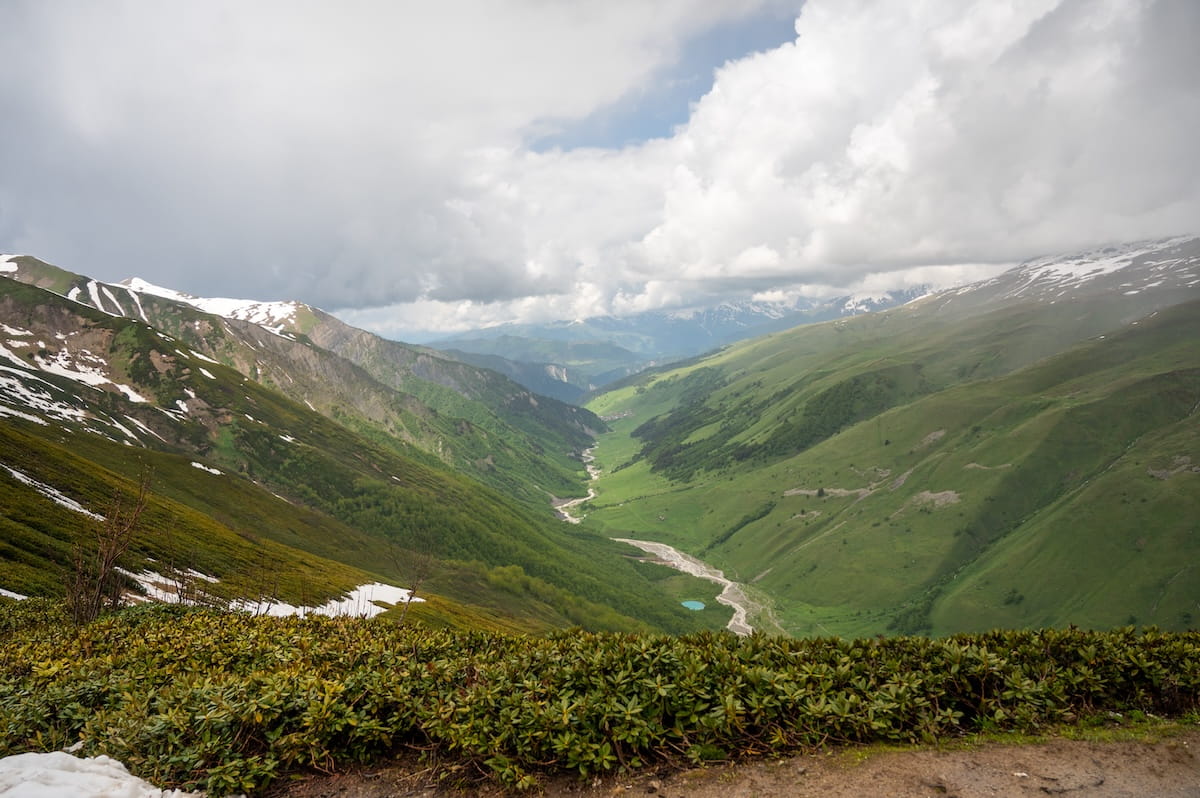 A high-angle view looking down into a deep, lush green V-shaped valley from Chkhunderi Pass, showcasing a winding river cutting through the valley floor, low alpine bushes in the foreground, and distant mountains under a heavy blanket of clouds in Svaneti, Georgia.