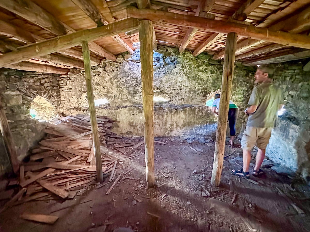 The rustic stone and wood interior of a 12th-century Svan defense tower in Chvabiani, showing wooden pillars supporting a timber ceiling, piles of old planks, and tourists looking through small, arched window cutouts.