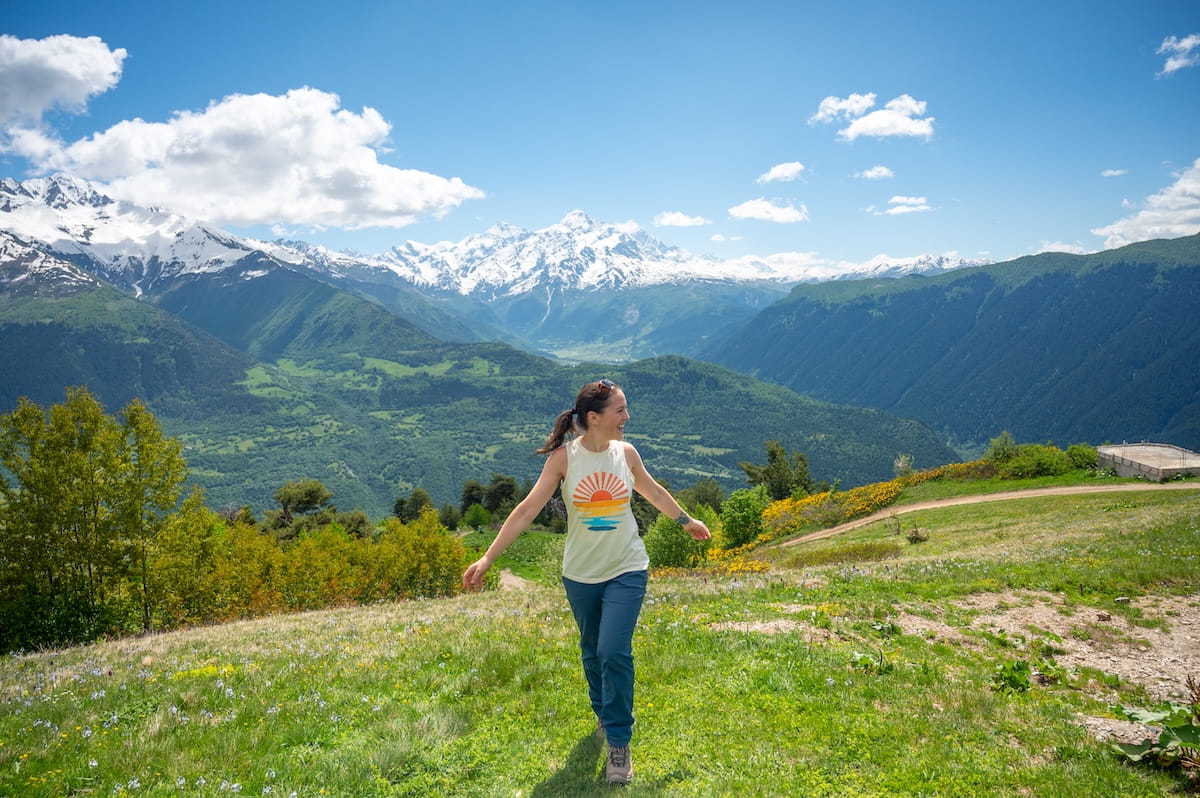 A happy female hiker walking down a grassy alpine ridge at the Cross Over Mestia viewpoint, showcasing expansive panoramic views of deep green valleys and towering, snow-capped Caucasus Mountain peaks in Svaneti, Georgia.