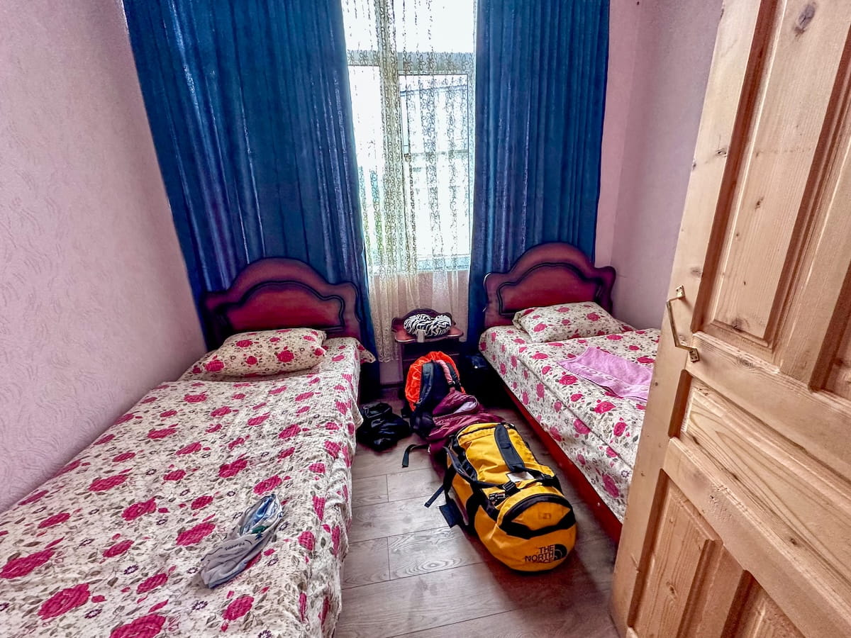 A small bedroom at Family Hotel Ucha in Iprali, featuring two twin beds with matching red and white floral duvets, a window with long blue curtains, and a yellow North Face duffel bag sitting on the wooden floor between the beds.