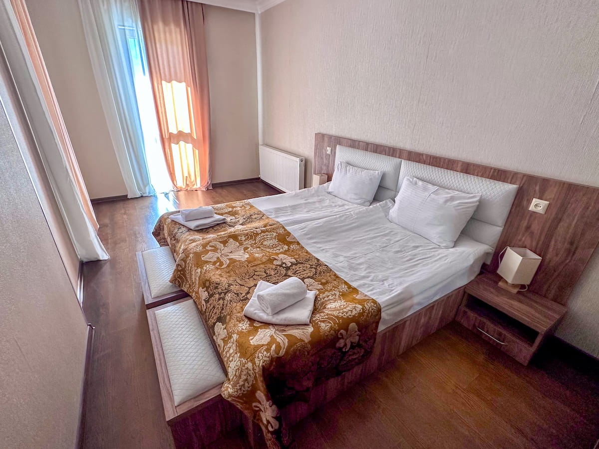A spacious hotel room at Hotel Shgedi Mestia, featuring a double bed with crisp white linens, a decorative gold patterned throw blanket, fresh folded towels, and hardwood flooring next to a balcony door with sheer curtains.