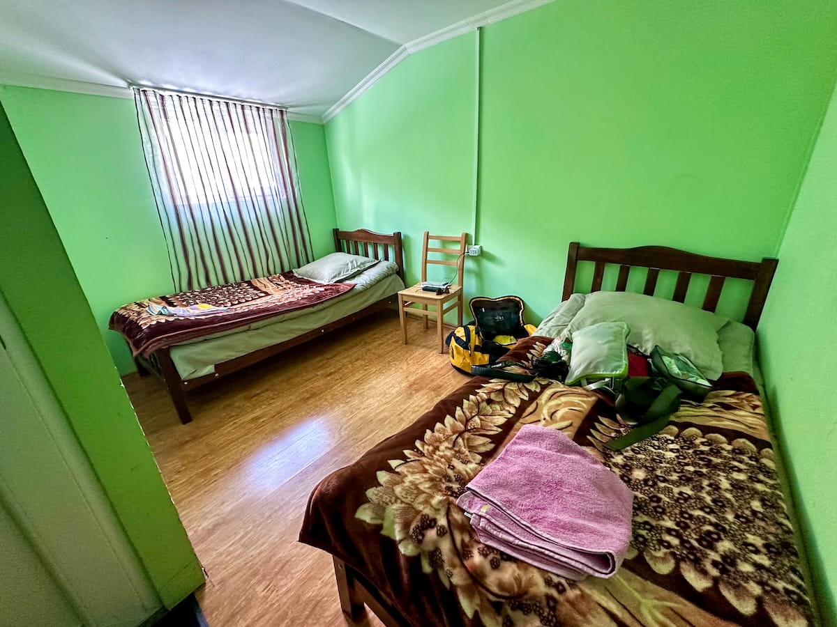 A simple hotel room at Hotel Tekla in Ushguli, featuring two single wooden beds with brown patterned plush blankets, bright lime green walls, a single wooden chair, and light-colored laminate flooring.