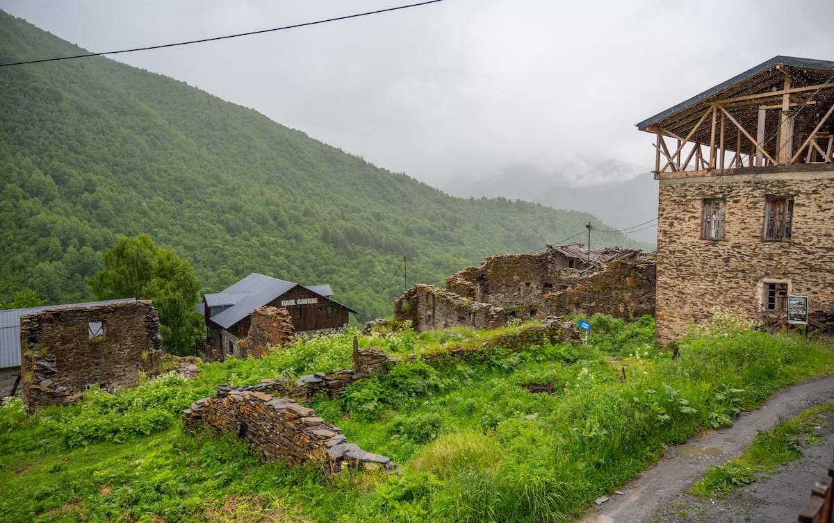 The stone ruins and crumbling walls of historic Khalde village on a rainy day, showcasing overgrown green grass and a gravel pathway next to newer guesthouses against a backdrop of mist-covered green mountains in Svaneti, Georgia.