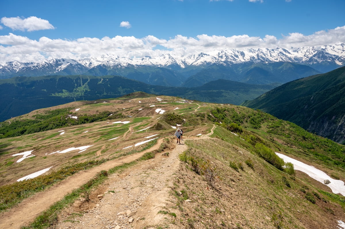 Hikers trekking along a wide dirt path up a steep mountain ridge on the Koruldi Lakes trail, with patches of snow on the green hillsides and a massive wall of snow-capped Caucasus peaks dominating the horizon in Svaneti, Georgia.