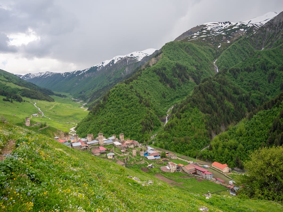 A high-angle panoramic view looking down at Adishi Village, showcasing its historic stone defensive towers and rustic homes nestled in a deep green valley beneath towering, snow-capped Caucasus Mountain ridges.