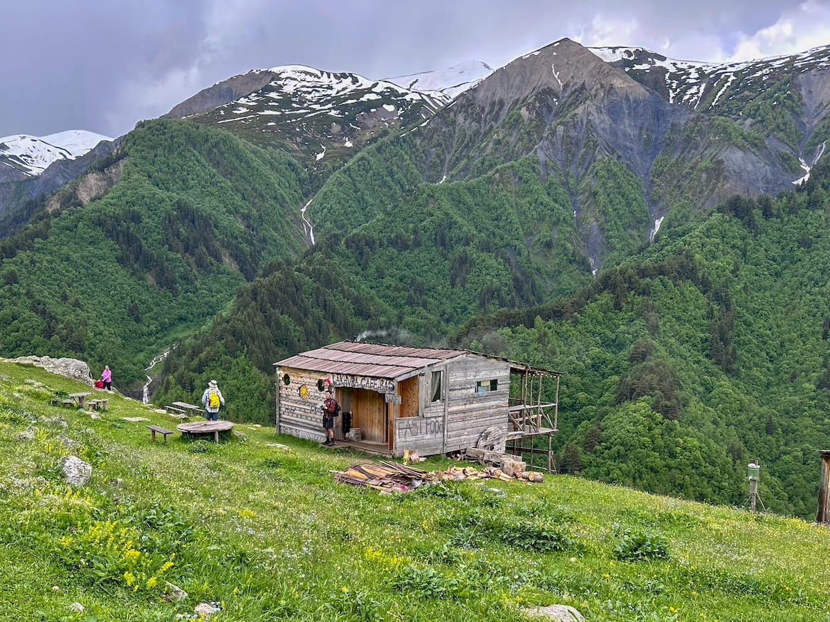 A rustic wooden cabin housing Lakhva Cafe Bar on a grassy alpine hillside in Adishi, with hikers resting outside against a spectacular backdrop of steep green mountainsides and distant snow-covered peaks in Svaneti, Georgia.