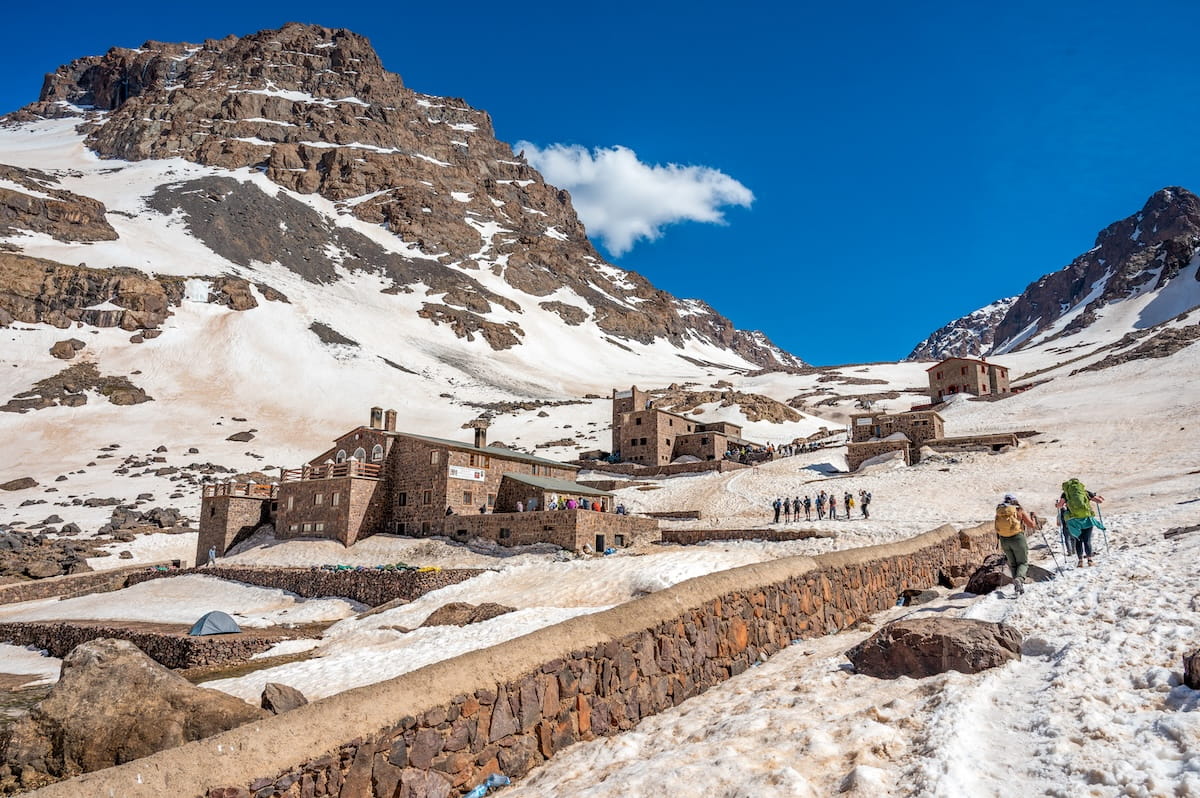 Hikers with backpacks walking along a snowy trail toward the stone buildings of the Toubkal Refuge in Morocco's High Atlas Mountains, surrounded by massive, snow-covered rocky peaks under a bright blue sky.