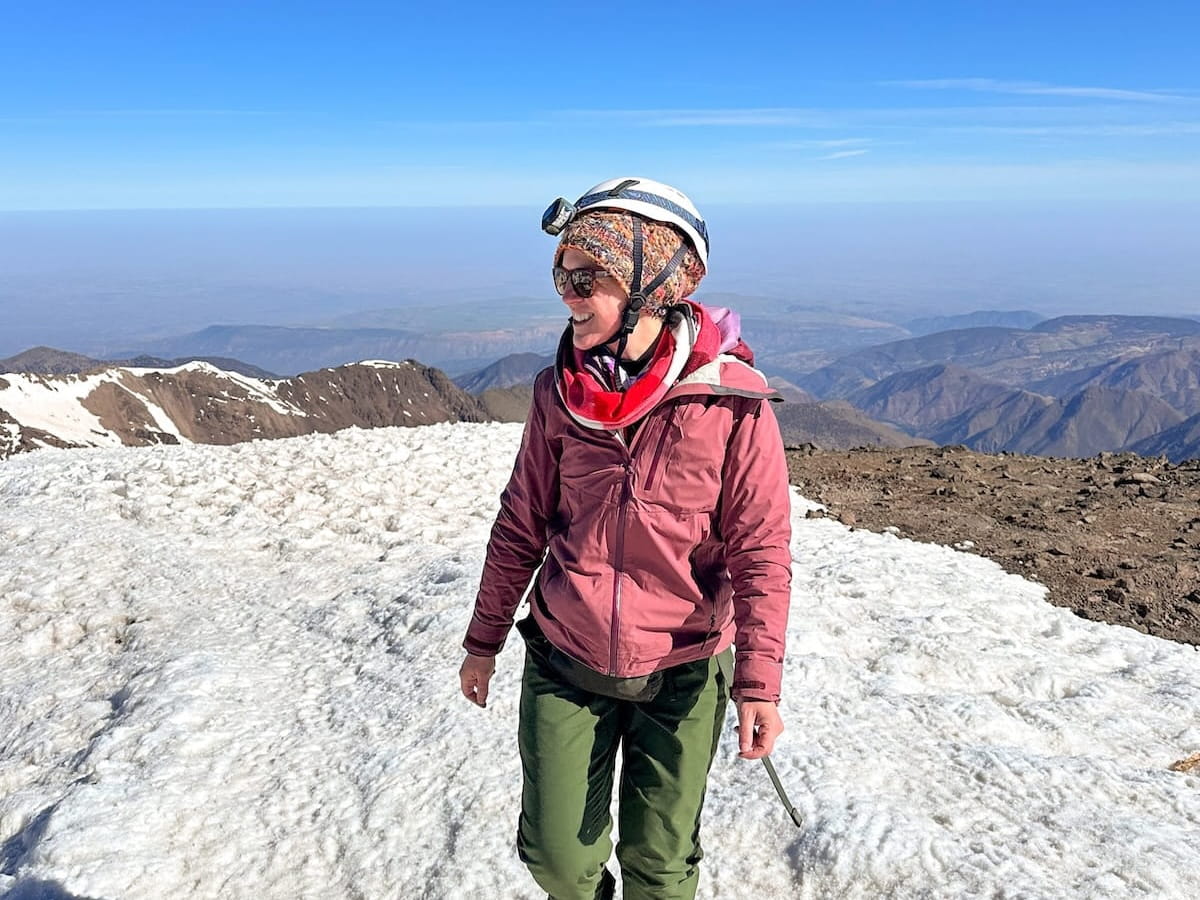 A female hiker walking on a snowy mountain ridge under a bright blue sky on the Mount Toubkal summit trail in Morocco. She is wearing green hiking pants, a pink winter jacket, a red scarf, a colorful knit hat, sunglasses, a climbing helmet with a headlamp, and crampons on her boots.