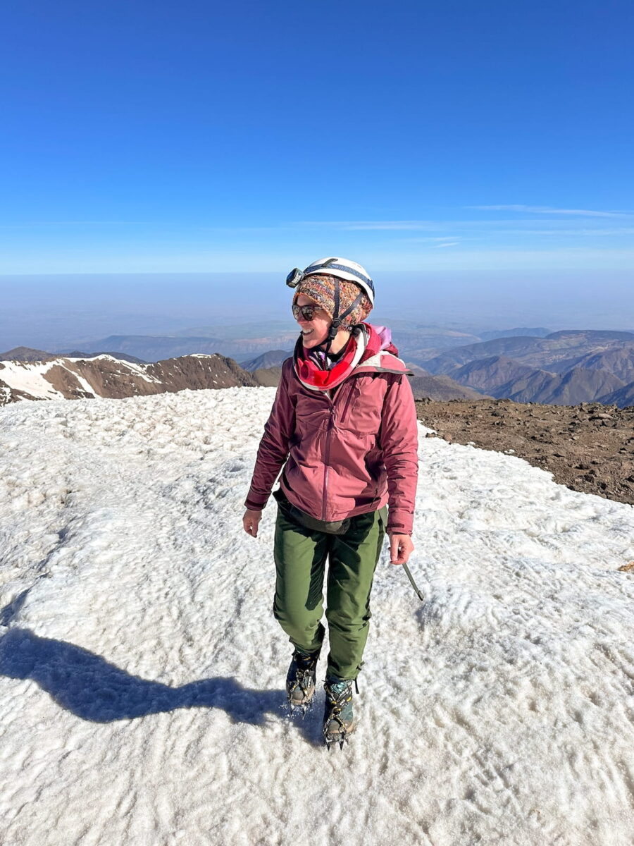 A female hiker walking on a snowy mountain ridge under a bright blue sky on the Mount Toubkal summit trail in Morocco. She is wearing green hiking pants, a pink winter jacket, a red scarf, a colorful knit hat, sunglasses, a climbing helmet with a headlamp, and crampons on her boots.