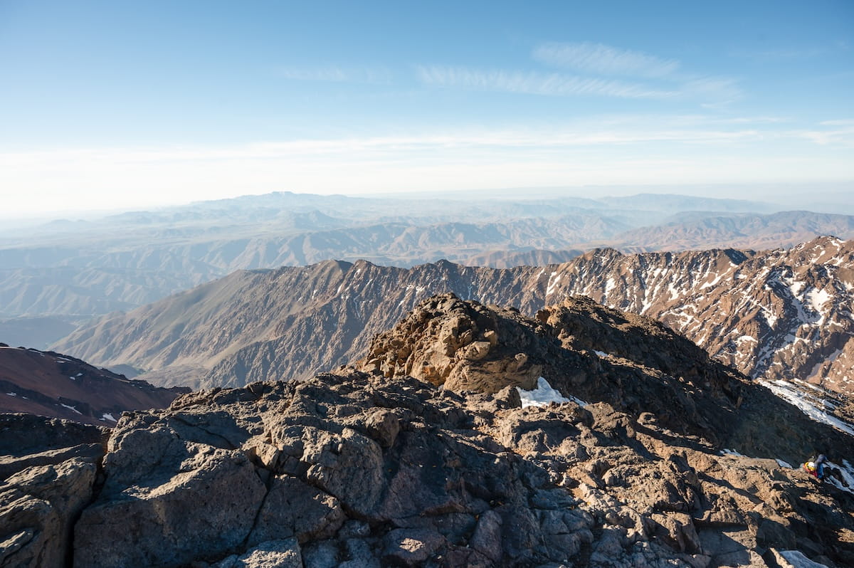 A breathtaking panoramic view looking out from the rocky summit of Mount Toubkal, Morocco. In the foreground are dark, rugged volcanic rock formations, while a vast sea of endless, dry mountain ridges and snow-dusted peaks of the High Atlas range stretches toward a hazy horizon under a pale blue sky.
