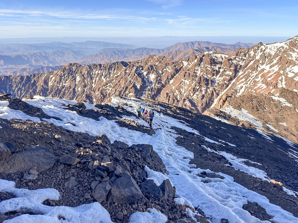 A small group of hikers ascending a steep, rocky trail covered in patches of snow on Mount Toubkal. The background shows panoramic views of the rugged, dry peaks of the High Atlas Mountains stretching out under a clear sky.