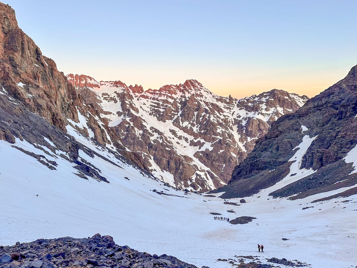 A wide scenic shot of a snowy mountain pass in the High Atlas Mountains of Morocco at sunrise. Distant groups of hikers are trekking across a vast snowy trail surrounded by towering, snow-covered rocky peaks under a soft orange and blue sky.