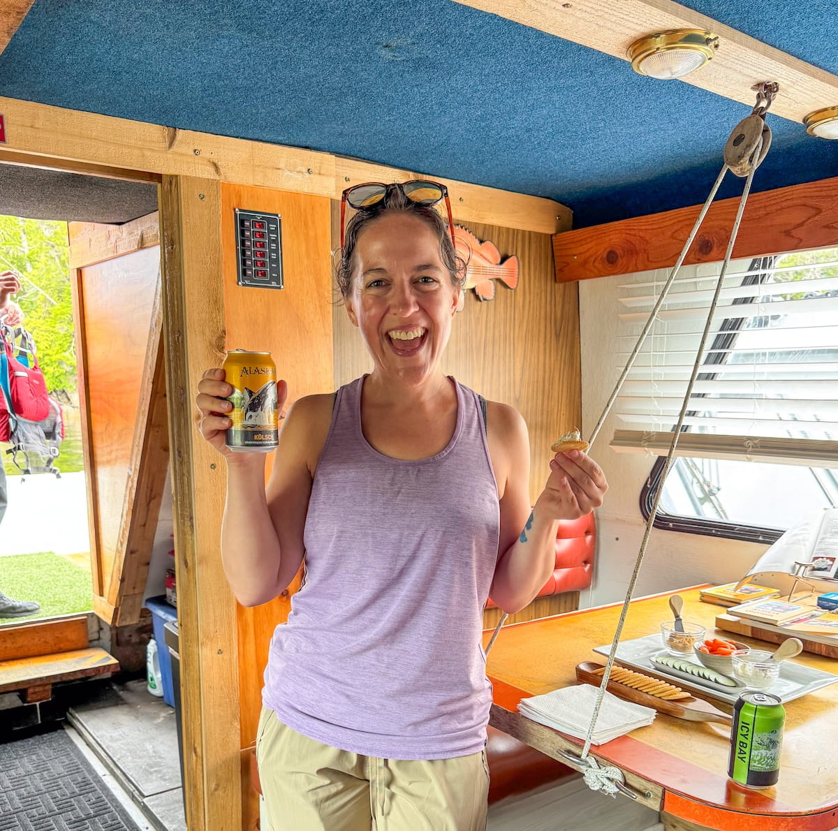 A smiling woman inside a cozy wood-paneled houseboat cabin holds a can of Alaskan Brewing Co. beer in one hand and a cracker topped with homemade salmon dip in the other, with a spread of snacks, crackers, and a can of Icy Bay beer visible on the table beside her.
