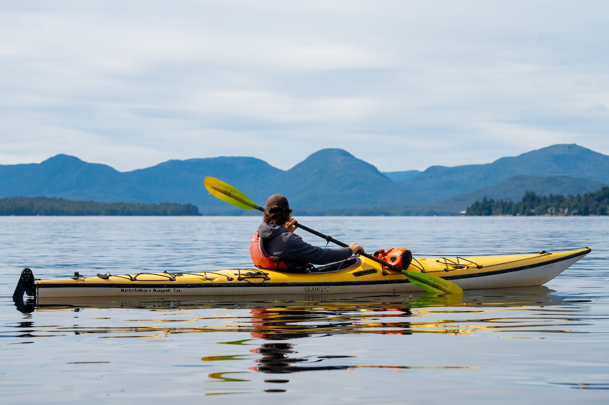 A tour guide seen from behind paddles a yellow and white sea kayak on calm, glassy water, with rolling blue mountain ranges and the forested shoreline of the Tongass National Forest under an overcast sky.