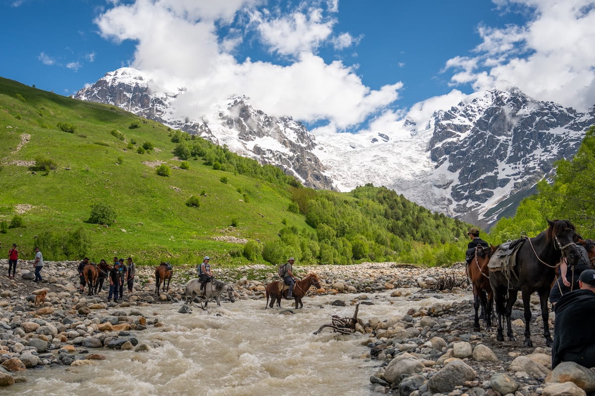 Hikers crossing the rushing, rocky Adishi River on horseback, with other trekkers waiting on the gravel banks under a spectacular view of the massive Adishi Glacier and snow-capped Caucasus peaks in Svaneti, Georgia.