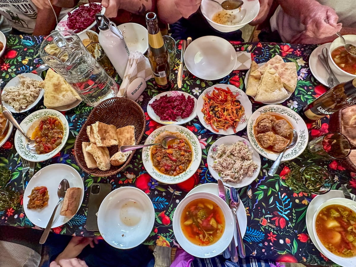An overhead view of a large, family-style Georgian food spread at a Svaneti guesthouse in Chvabiani, featuring multiple plates of vegetable soups, shredded meat salad, red beetroot salad, triangles of traditional khachapuri cheese bread, and dips on a colorful floral tablecloth.