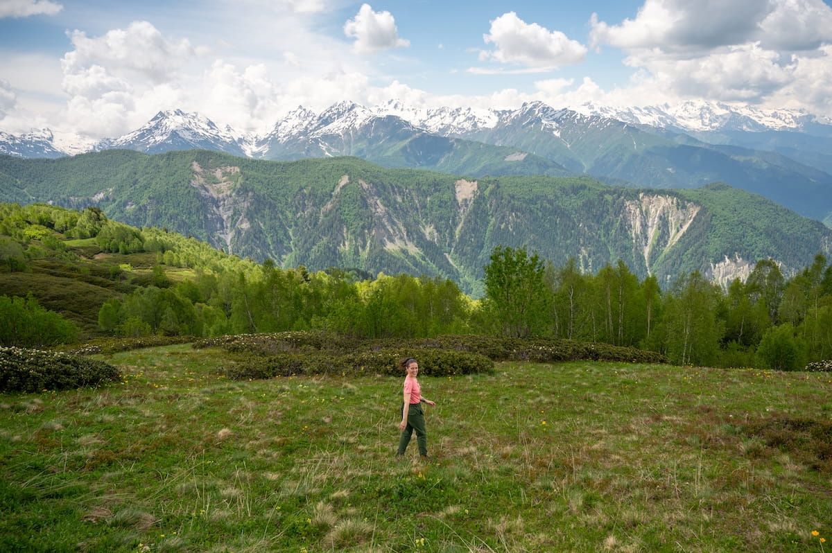 A female hiker walking through a vast green alpine meadow near Tetnuldi Ski Resort, looking back toward the camera with a breathtaking backdrop of forested hills and the snow-capped Caucasus Mountain peaks stretching across the horizon in Svaneti, Georgia.
