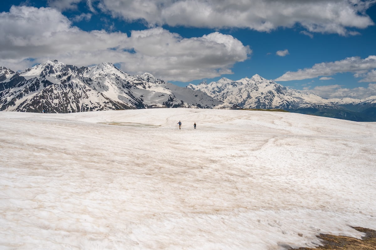 Two distant hikers using trekking poles to cross a massive alpine snowfield on the trail to Koruldi Lakes near Mestia, surrounded by the towering, jagged, snow-capped peaks of the Caucasus Mountains under a partly cloudy blue sky in Svaneti, Georgia.