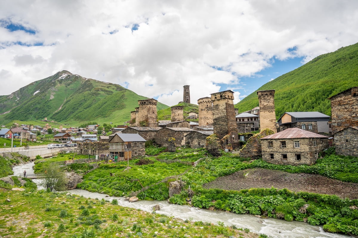 A scenic view of Chazhashi village in the Ushguli community, showcasing its ancient, UNESCO-listed stone defensive towers and rustic stone houses built along a rushing river, surrounded by green mountains under a cloudy sky in Svaneti, Georgia.