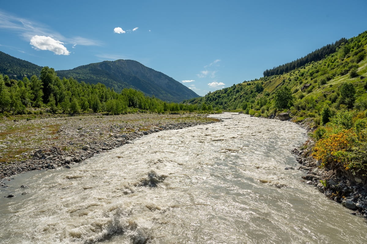 A wide, fast-flowing river with churning silty water rushing through a rocky gravel valley, lined with dense green forests and rolling hillsides under a bright blue sky in Svaneti, Georgia.