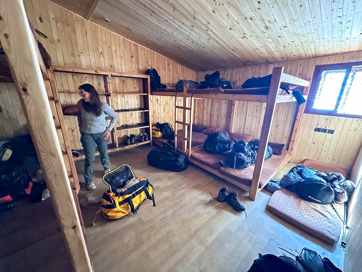 Inside a rustic, wood-paneled communal dorm room at Les Mouflons Refuge in the Toubkal Refuge complex, featuring multi-tiered wooden bunk beds with brown mattresses and pillows, open wooden storage shelves, and a hiker standing next to duffel bags and backpacks on the floor.