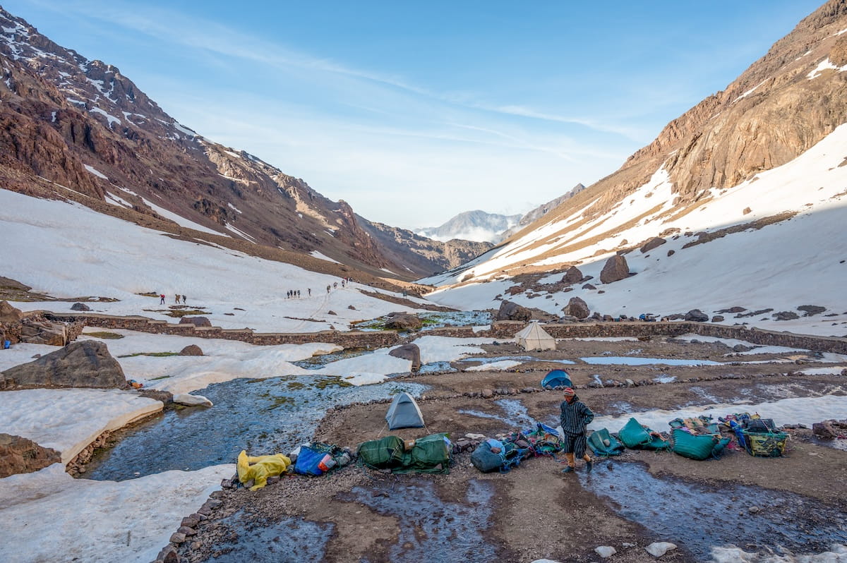 A scenic mountain valley view from the Toubkal Refuge in Morocco, showing small camping tents pitched on rocky ground next to a flowing stream, with a line of tiny hikers trekking across distant snow-covered slopes between massive mountain ridges.
