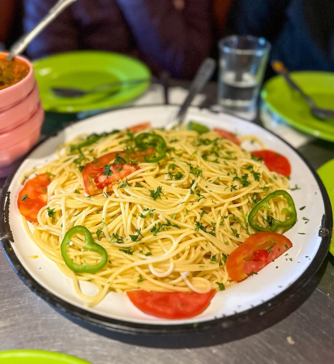 A large plate of spaghetti pasta topped with sliced green peppers, tomatoes, and fresh chopped herbs, served family-style on a dining table at the Toubkal Refuge during a guided mountain trek in Morocco.munal dorm room at Les Mouflons Refuge in the Toubkal Refuge complex, featuring multi-tiered wooden bunk beds with brown mattresses and pillows, open wooden storage shelves, and a hiker standing next to duffel bags and backpacks on the floor.