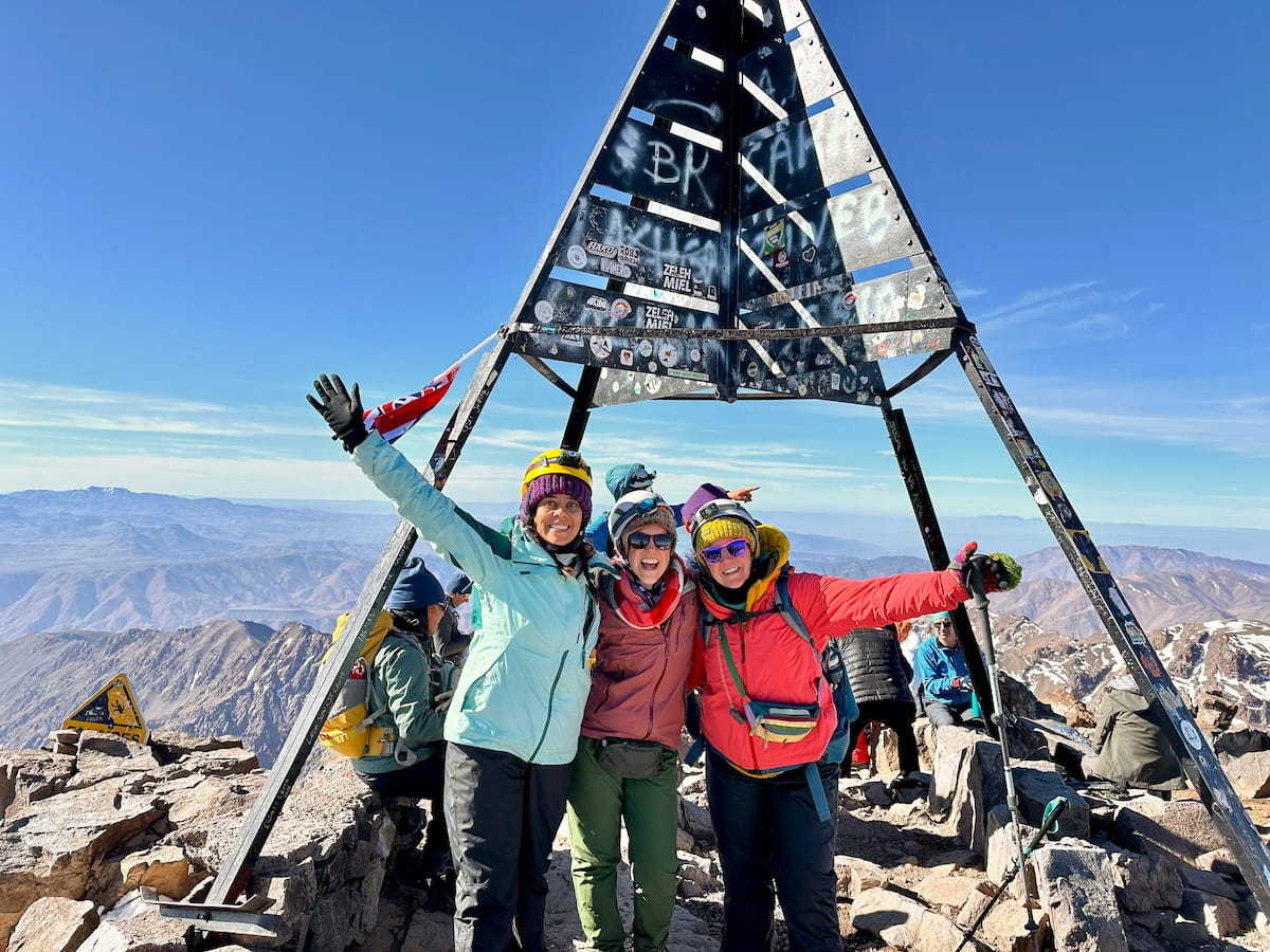 Three smiling female hikers in colorful winter jackets and sunglasses posing arm-in-arm beneath the iconic black metal triangular summit structure on Mount Toubkal, with endless mountain peaks under a bright blue sky.