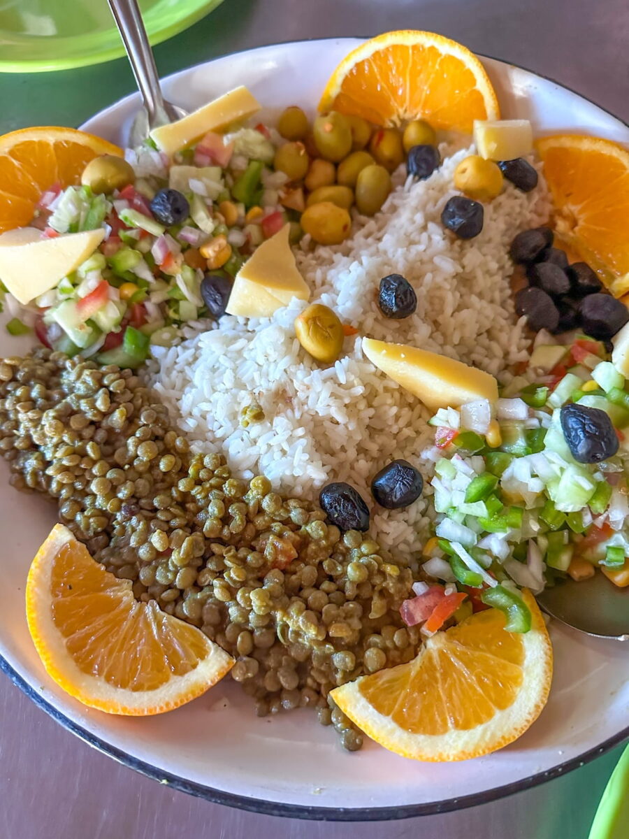 A large white plate filled with a traditional Moroccan hiking lunch of white rice, cooked lentils, diced cucumber and tomato salad, green and black olives, triangles of cheese, and orange slices arranged around the rim.
