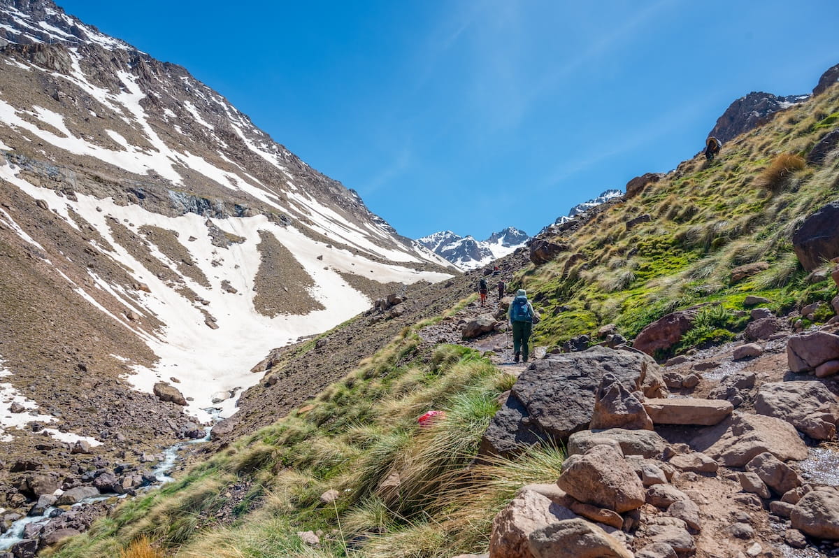 Hikers walking up a rocky dirt trail along a grassy mountain slope on the route from Imlil to the Toubkal Refuge. To the left is a steep mountain face covered in melting snow with a small stream running through the valley floor.