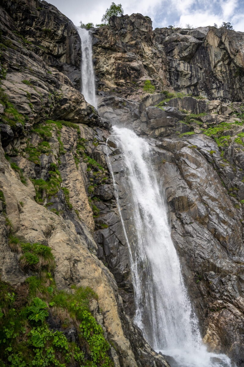 A low-angle shot of the powerful Ushba (Shdugra) Waterfall cascading down a massive, steep rocky cliff face in the Svaneti region of Georgia.
