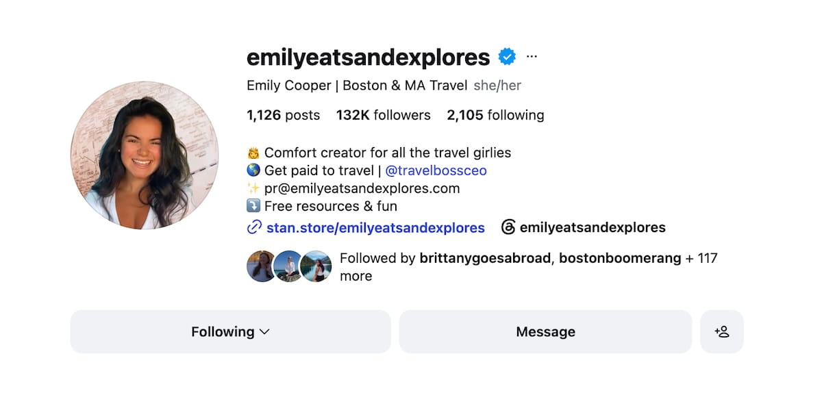 A screenshot of an Instagram profile page for the verified account emilyeatsandexplores. The circular profile picture features a smiling woman with dark hair in front of a world map. The profile text reads "Emily Cooper | Boston & MA Travel she/her" with stats showing 1,126 posts, 132K followers, and 2,105 following. The bio includes bulleted lines: "Comfort creator for all the travel girlies," "Get paid to travel | @travelbossceo," contact email "pr@emilyeatsandexplores.com," "Free resources & fun," and a link to "stan.store/emilyeatsandexplores."