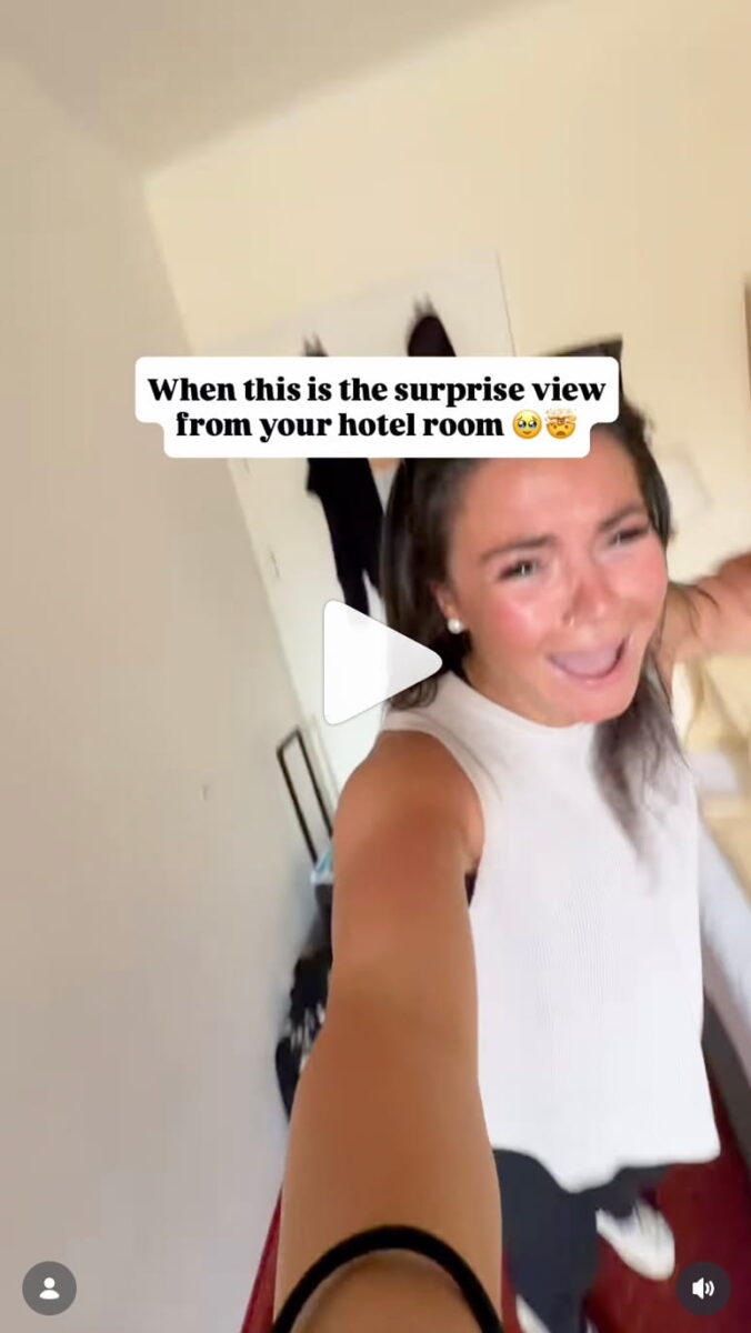 A vertical screenshot of an Instagram Reel video showing a dynamic, handheld self-recorded frame. A woman in a sleeveless white top holds the camera at a high angle while walking backward through a hotel room with an excited, wide-mouthed expression. A white text overlay near the top of the frame reads "When this is the surprise view from your hotel room" followed by an emotional face emoji and a star-struck emoji. A white play icon sits in the center of the screen.