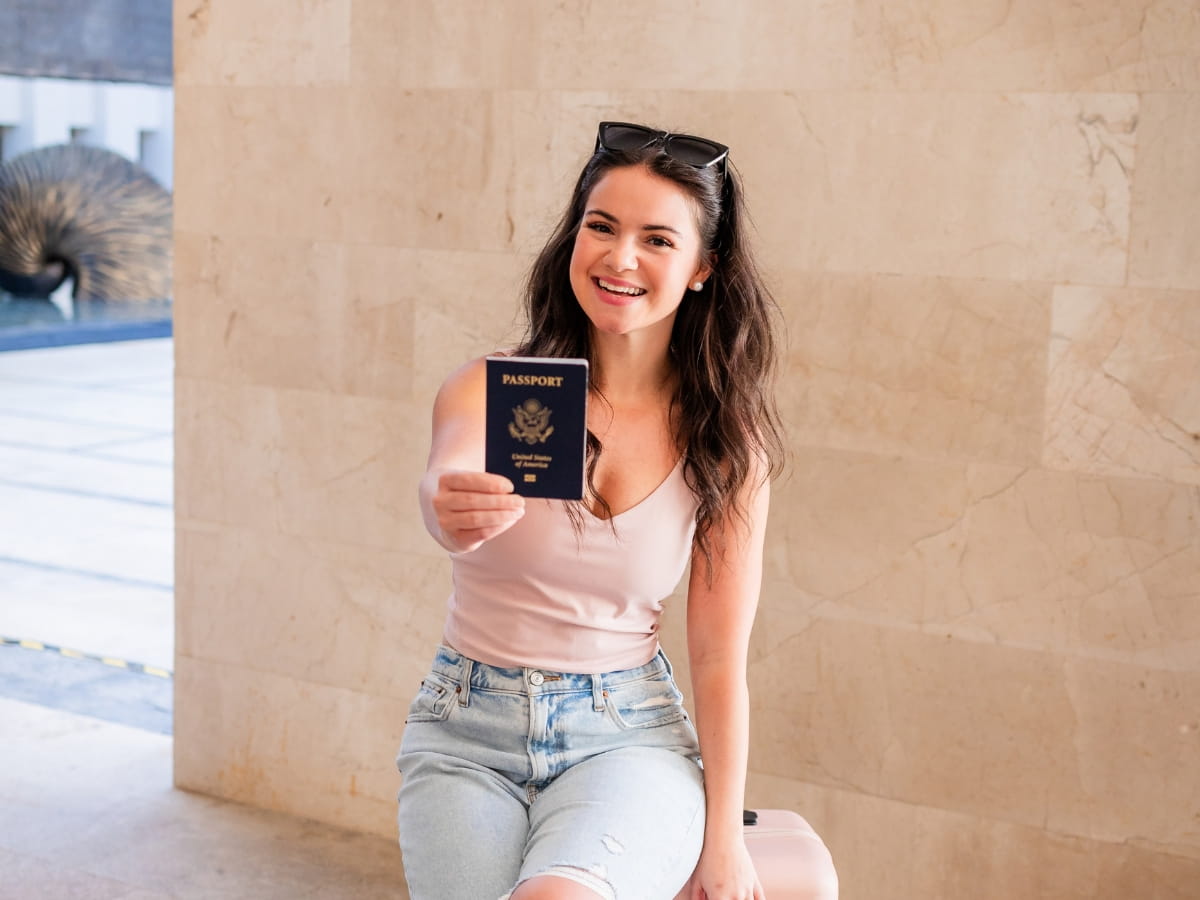 A medium, eye-level shot of a smiling travel creator sitting on a light pink hardshell suitcase, holding up a blue United States passport directly toward the camera. She has long, dark wavy hair with sunglasses resting on her head, wearing a light pink tank top and light-wash ripped jeans. She is positioned against a smooth, cream-colored stone wall in a bright, modern terminal or hotel lobby.