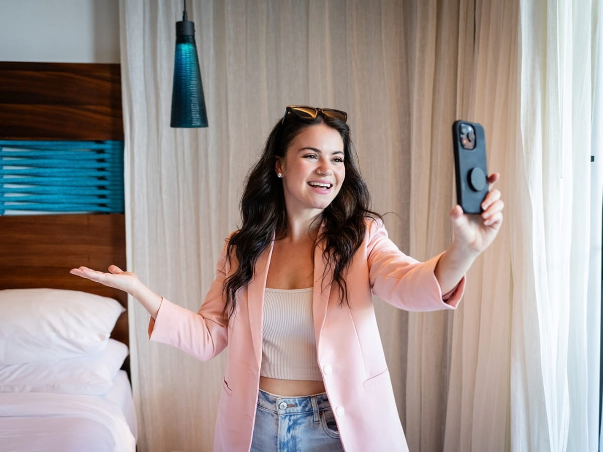 A female travel creator smiling while holding a smartphone up to film Instagram content in a stylish hotel room. She has wavy dark hair with sunglasses resting on her head, and she is wearing a light pink blazer, a white ribbed crop top, and blue jeans. Her left hand gestures welcomingly toward a neatly made bed in the background, set against sheer white curtains and a modern blue-accented wooden headboard.