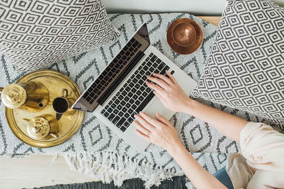 A person working on a laptop from home, seated on a patterned sofa with a cup of coffee on a tray beside them.