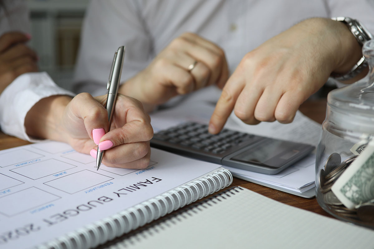 Couple planning a family budget together at a table, with one person writing in a planner while the other uses a calculator; coins and cash are visible in a glass jar nearby.