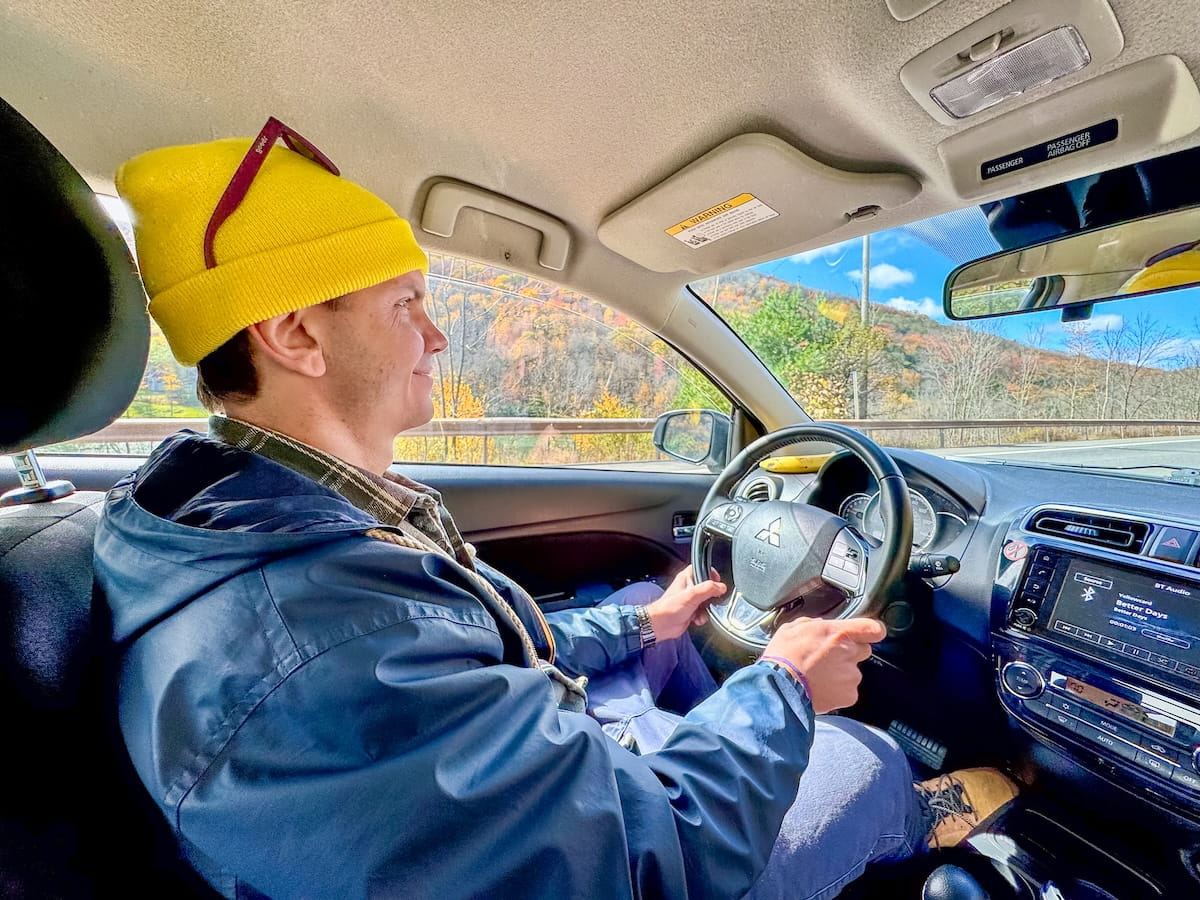 An interior car photo taken from the back seat showing a male driver wearing a bright yellow beanie, sunglasses resting on his hat, a dark jacket, and blue jeans, steering a Mitsubishi along a scenic highway during a Catskills fall road trip. Outside the passenger-side window and windshield, autumn mountainside foliage in shades of orange and yellow glows under a bright blue sky.