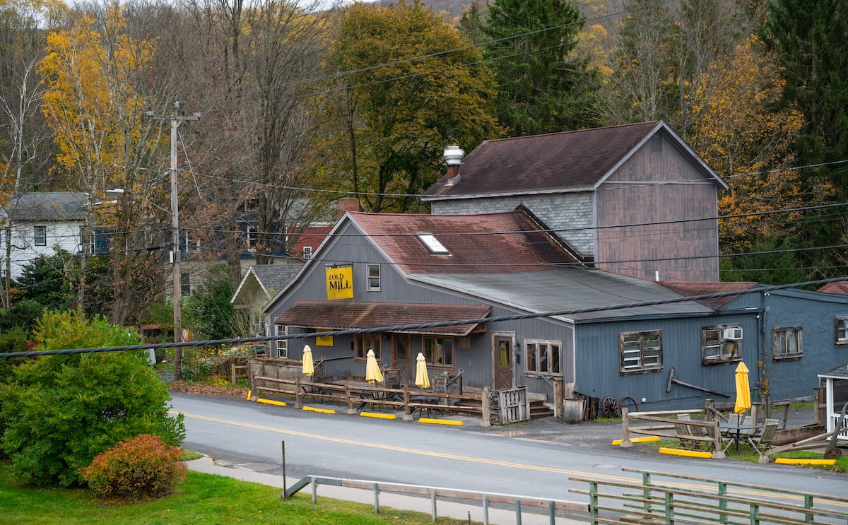A high-angle exterior shot of The Old Mill Restaurant in Roxbury, NY, located along a two-lane country road in the Catskills. The rustic grey wooden building features a brown shingled roof, a yellow "The Old Mill" sign, and an outdoor patio area with wooden fencing and closed yellow umbrellas. In the background, lush forest trees show early autumn colors, and nearby houses nestle along the scenic roadside.