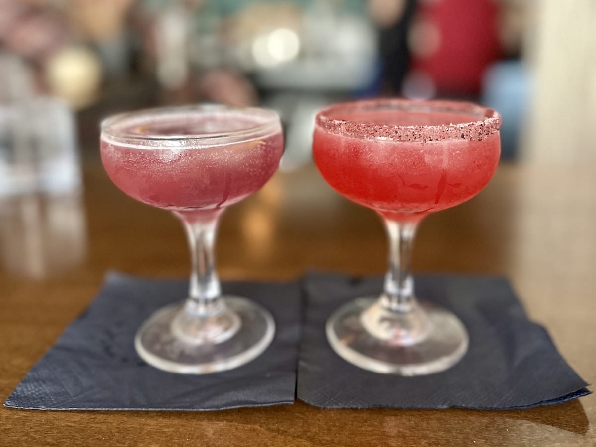 A close-up shot of two craft cocktails served in coupe glasses resting on dark paper cocktail napkins on a wooden table at The Print House in Fleischmanns, NY. The cocktail on the left is a soft translucent pink with a citrus twist, while the cocktail on the right is a vibrant red with a dark spiced rim.
